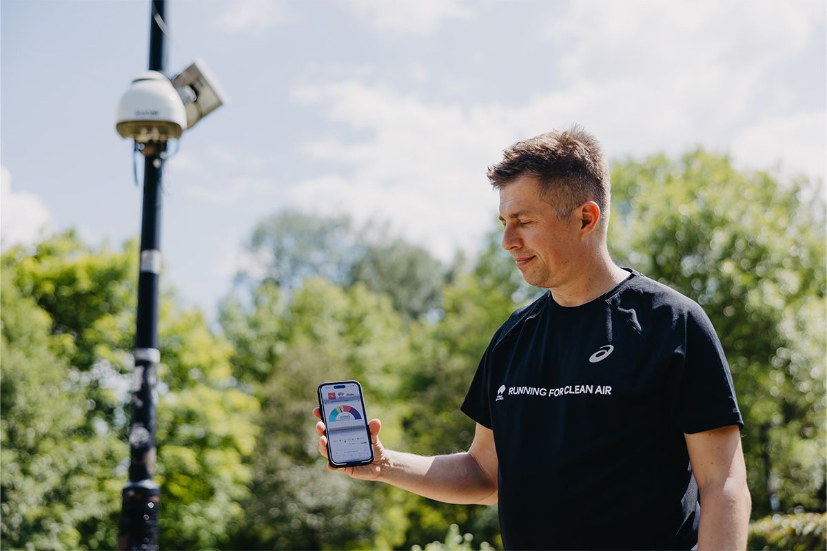 Un miembro del equipo del proyecto Running for Clean Air junto a una estación Kunak AIR Pro instalada en una farola en Varsovia y sosteniendo un móvil donde se visualizan los datos de calidad del aire en tiempo real - Kunak
